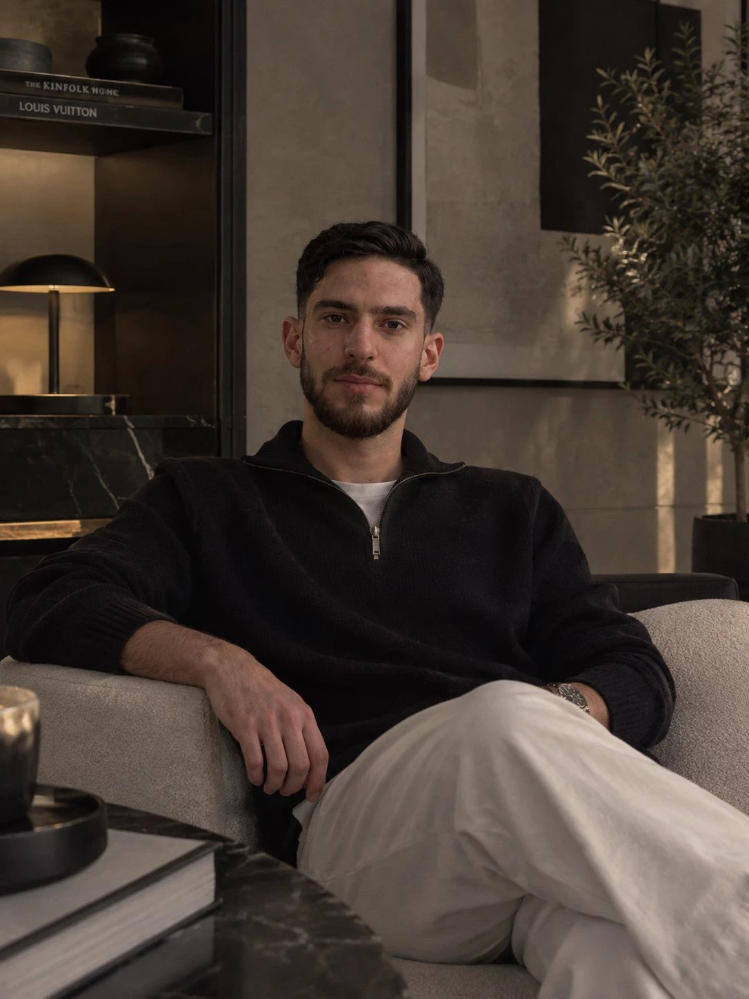 Mohammed in a black half-zip sweater and white trousers, relaxed on a linen armchair